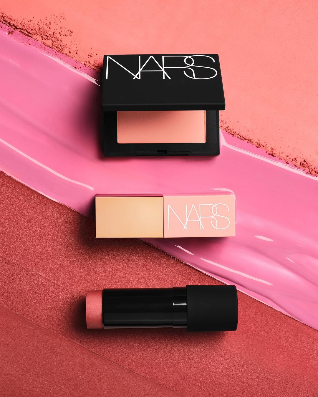 NARS blush and lip products arranged on a gradient pink surface, showcasing makeup and cosmetic products in a studio setting.