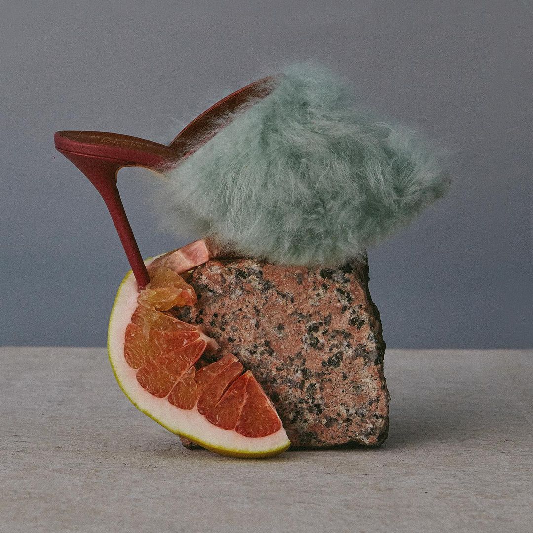 Still life of a red heeled shoe with light blue fur, grapefruit wedge, and textured brown rock against a graduated blue background.