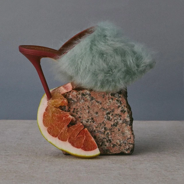 Still life of a red heeled shoe with light blue fur, grapefruit wedge, and textured brown rock against a graduated blue background.