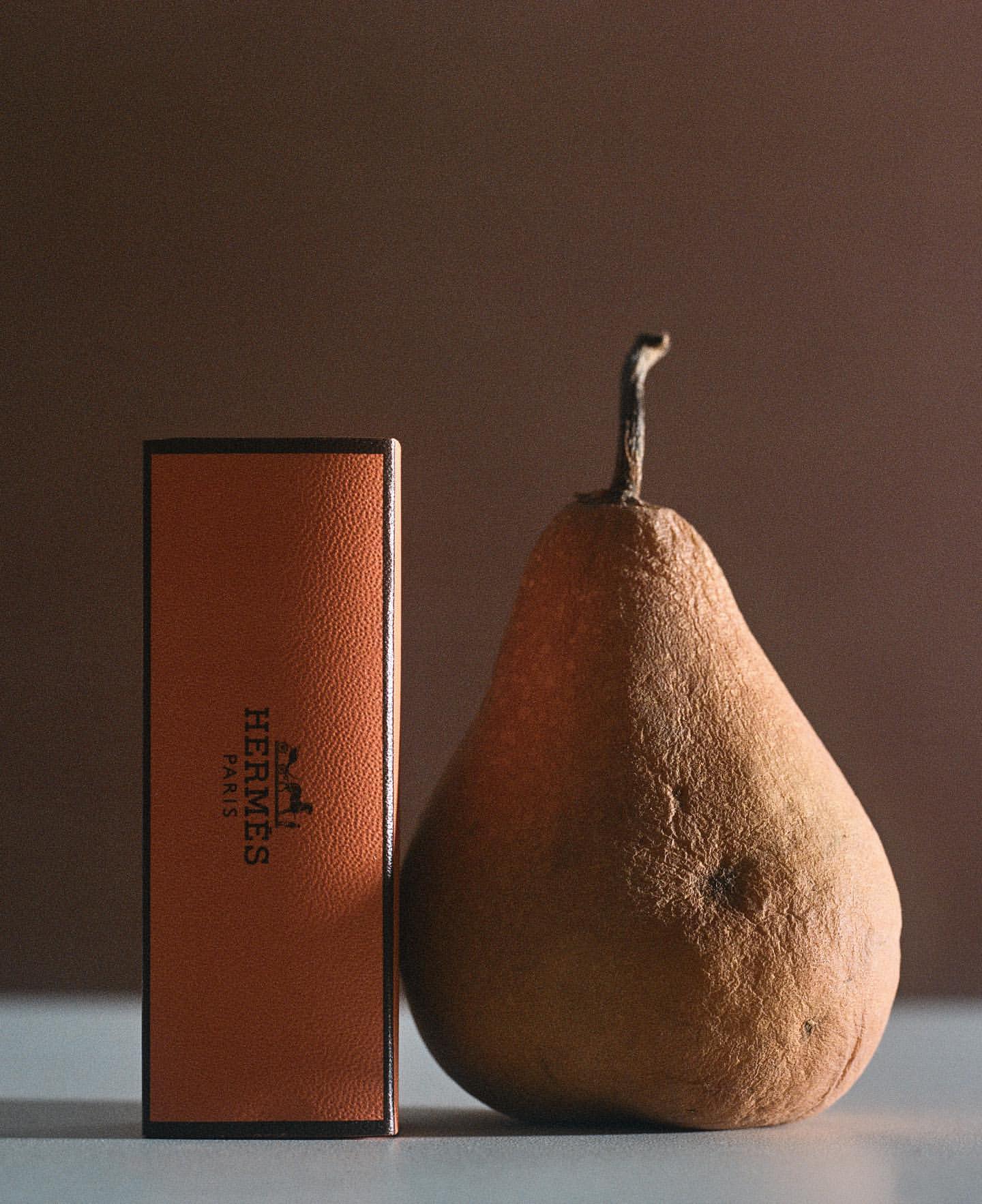 Still life shot of an oblong brown pear with stem next to a tall narrow perfume box labeled HERMES in a dimly lit setting.
