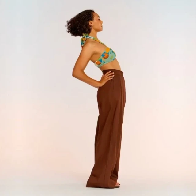 A woman in a halter top and wide-legged pants stands indoors with her hands on her hips, arching her back in a fashion pose.