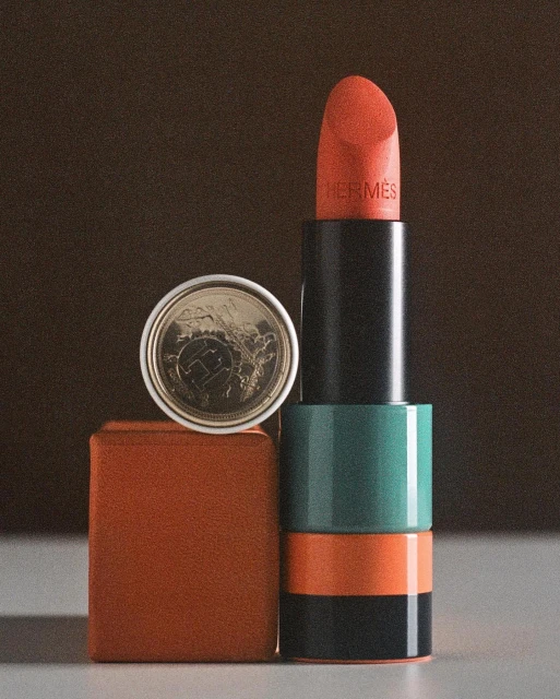 Still life shot of Hermès lipstick in red orange beside a euro cent coin on a brown cube against a blurred brown background.