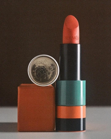 Still life shot of Hermès lipstick in red orange beside a euro cent coin on a brown cube against a blurred brown background.