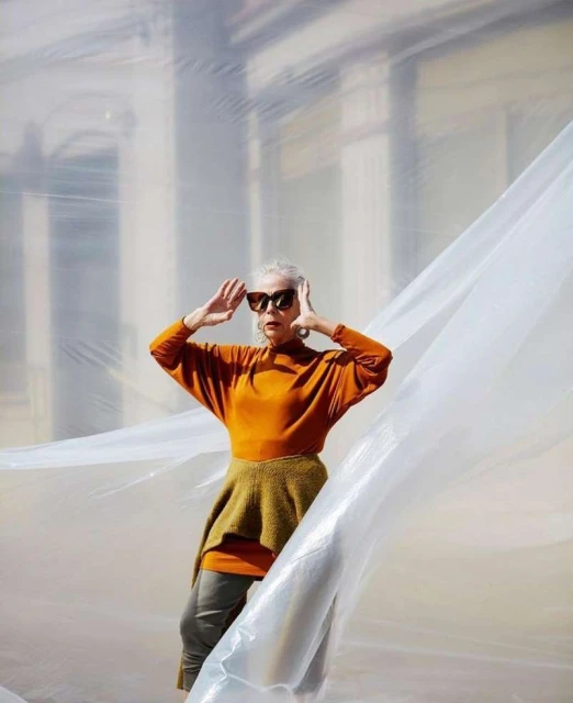 Elderly woman in orange top posing in front of a building with large plastic sheet in the foreground, wearing sunglasses and earrings.