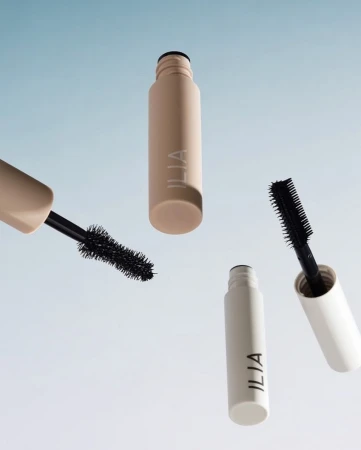 Two tubes of Ilia brand mascara, one beige and one white, float against a light blue and white gradient background. Open tube detail.