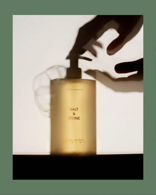 Cosmetic bottle with pump dispenser under shadow of hand. Salt & Stone product shot. Neutral-toned beauty product in studio setting.