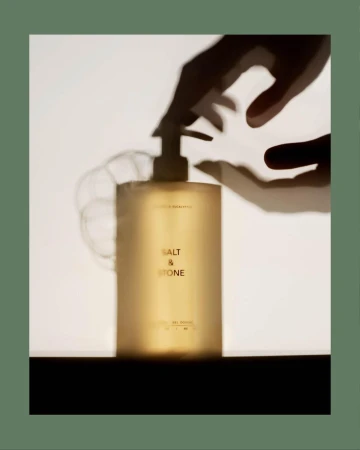Cosmetic bottle with pump dispenser under shadow of hand. Salt & Stone product shot. Neutral-toned beauty product in studio setting.