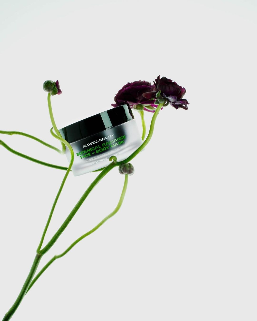 Elevated product shot of Allwell Beauty Botanical Radiance Face + Body Mask alongside delicate purple flowers on a clean white background.