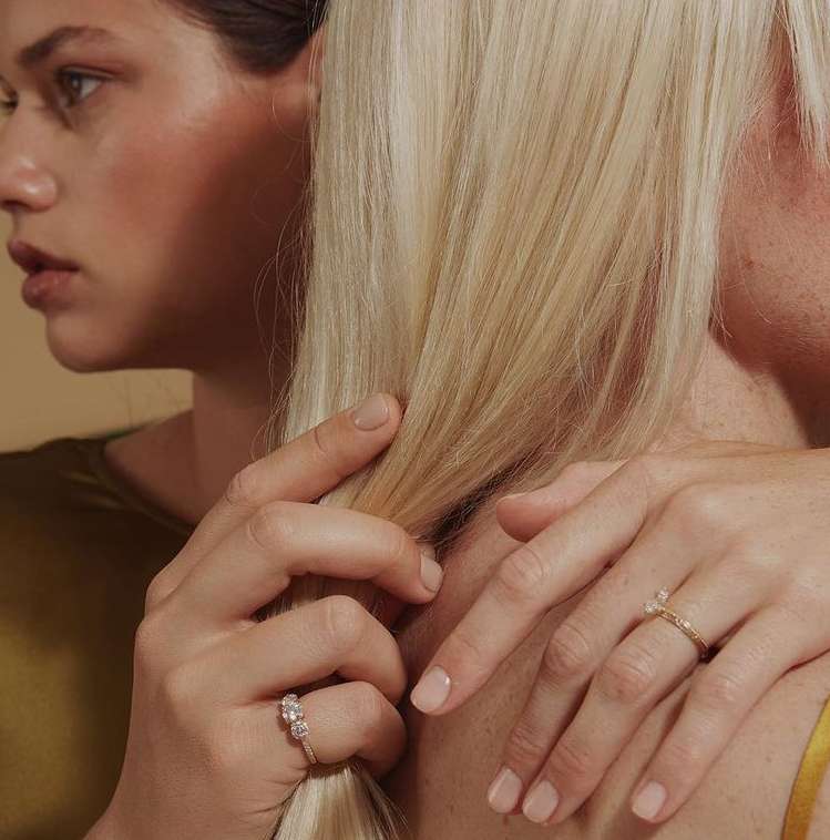 Close-up studio shot of two women showcasing engagement rings with neutral manicures and blonde hair against a beige backdrop.