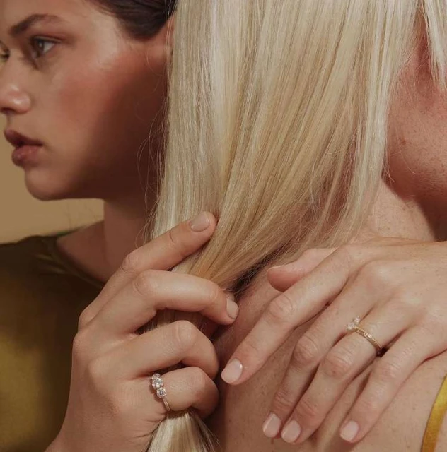 Close-up studio shot of two women showcasing engagement rings with neutral manicures and blonde hair against a beige backdrop.