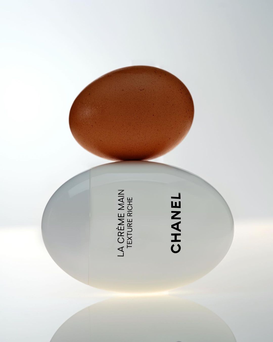 Egg sits atop a white, ovate bottle of Chanel La Crème Main Texture Riche cream in a studio setting with a gradient background.