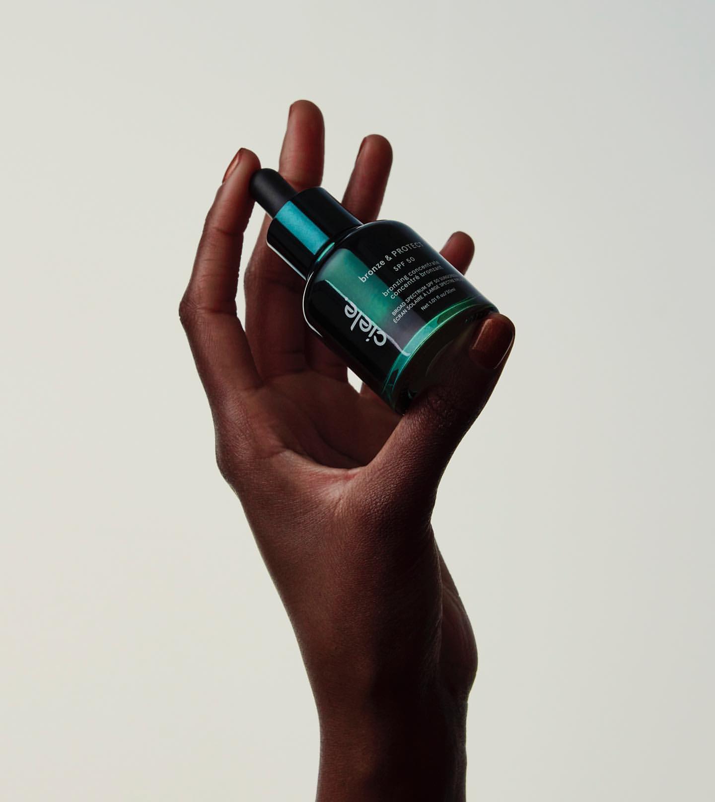 Dark-skinned hand holds a dark green glass bottle with a black cap against a pale background, showcasing beauty and skincare.