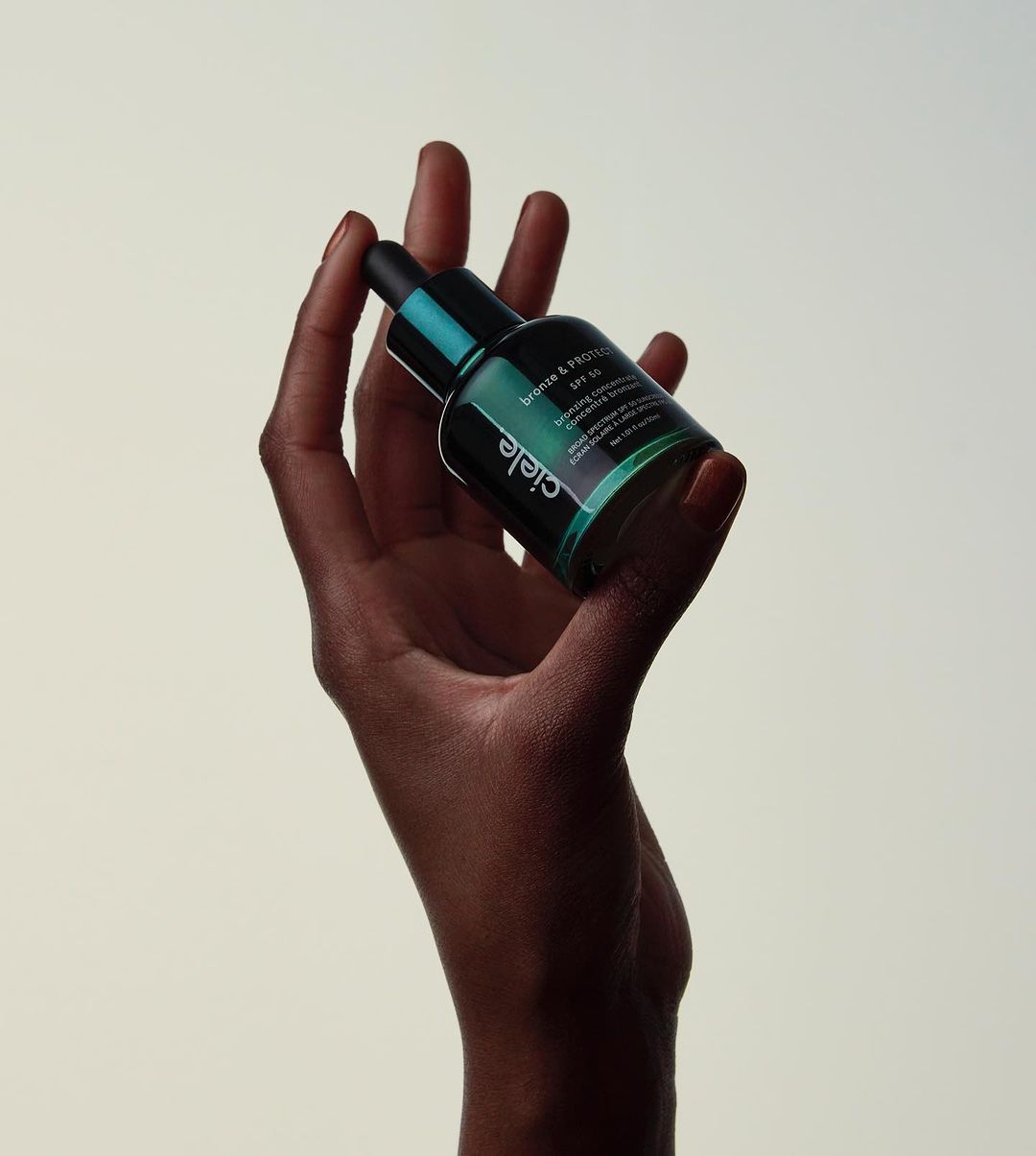 Ciele skincare bronze and protect SPF 50 serum bottle held by an adult female hand against a cream-colored background.
