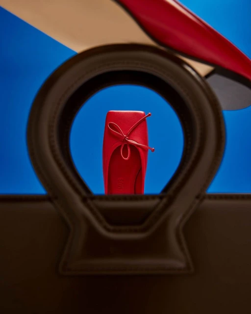 Red ballet flat and patent pump shoes are seen through the handle of a brown leather handbag against a two-tone blue backdrop.