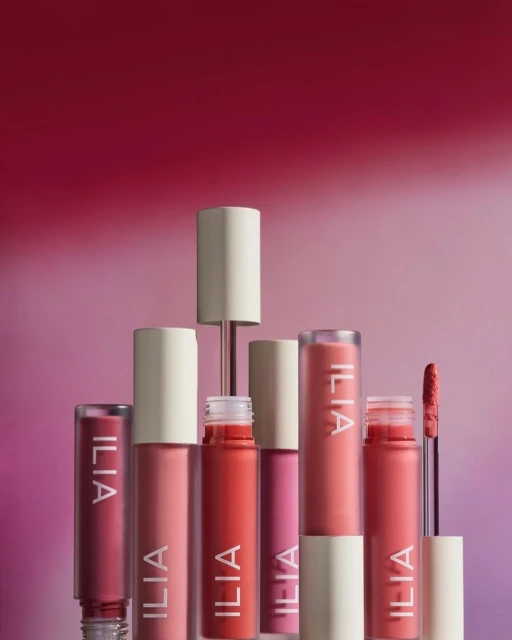 A collection of colorful Ilia lip gloss tubes are displayed in a studio setting with pink and red gradient background, showcasing beauty products.