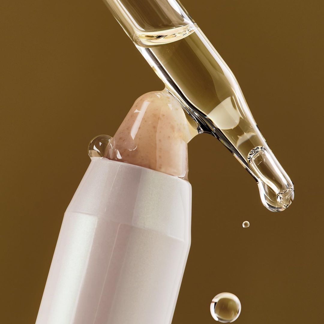 Serum dropping from a glass dropper onto a cosmetic product against a tan background, conveying beauty and skincare.