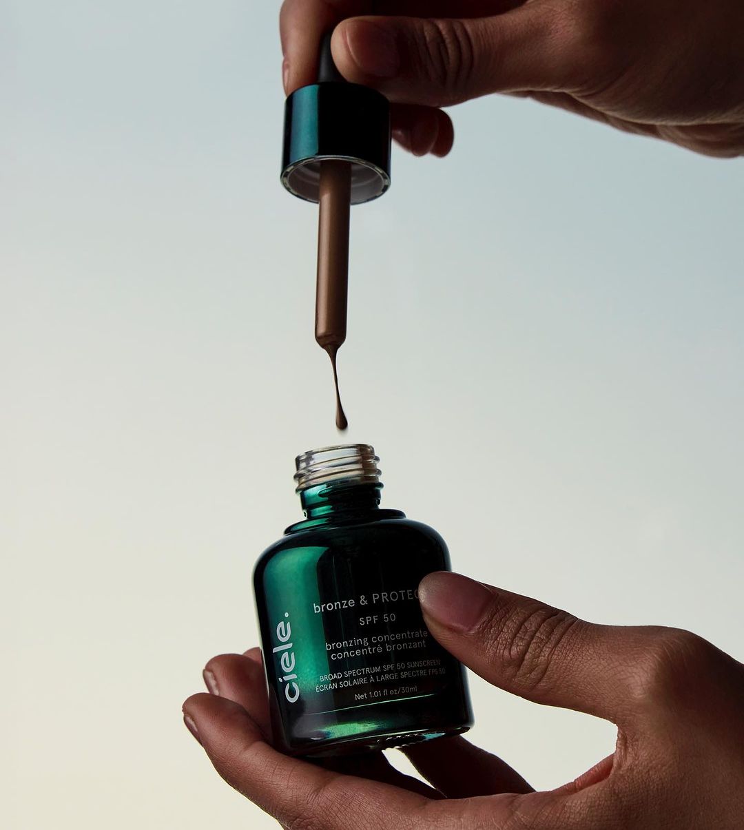 Close-up shot of hands holding a bottle of bronze and protein SPF 50 with a dropper, against a light background.