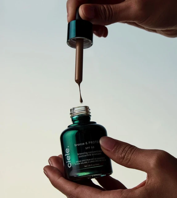 Close-up shot of hands holding a bottle of bronze and protein SPF 50 with a dropper, against a light background.