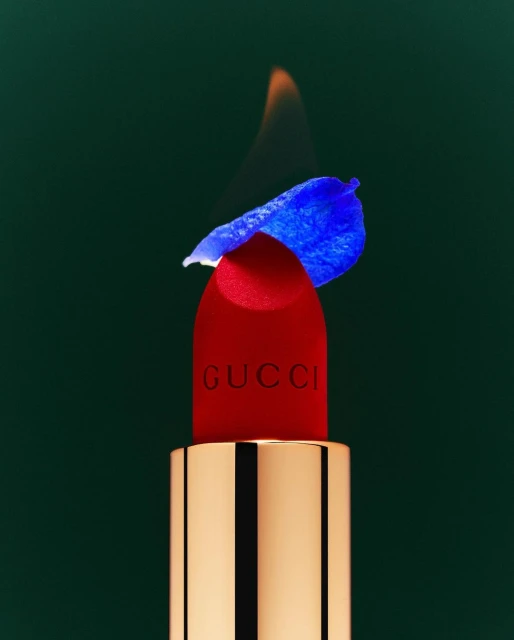 Gucci lipstick with blue flower petal and a flame on top, against a dark green backdrop in a beauty and fashion still life photograph.