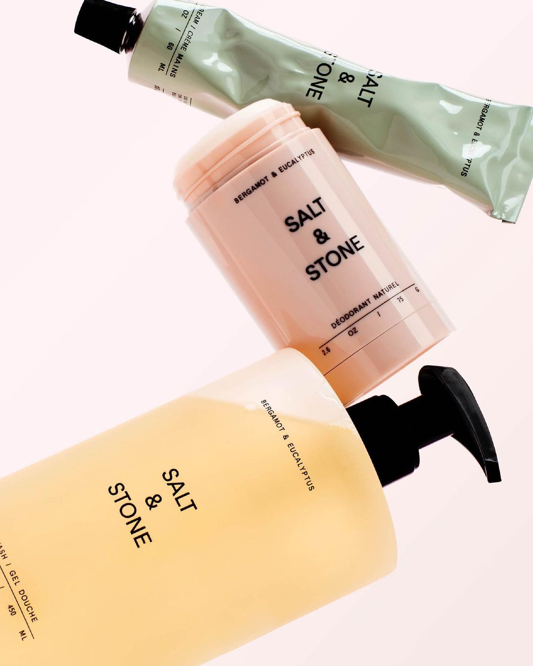 Close-up of Salt & Stone brand beauty products including hand cream, deodorant, and body wash, against a light pink studio backdrop.