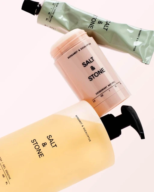 Close-up of Salt & Stone brand beauty products including hand cream, deodorant, and body wash, against a light pink studio backdrop.