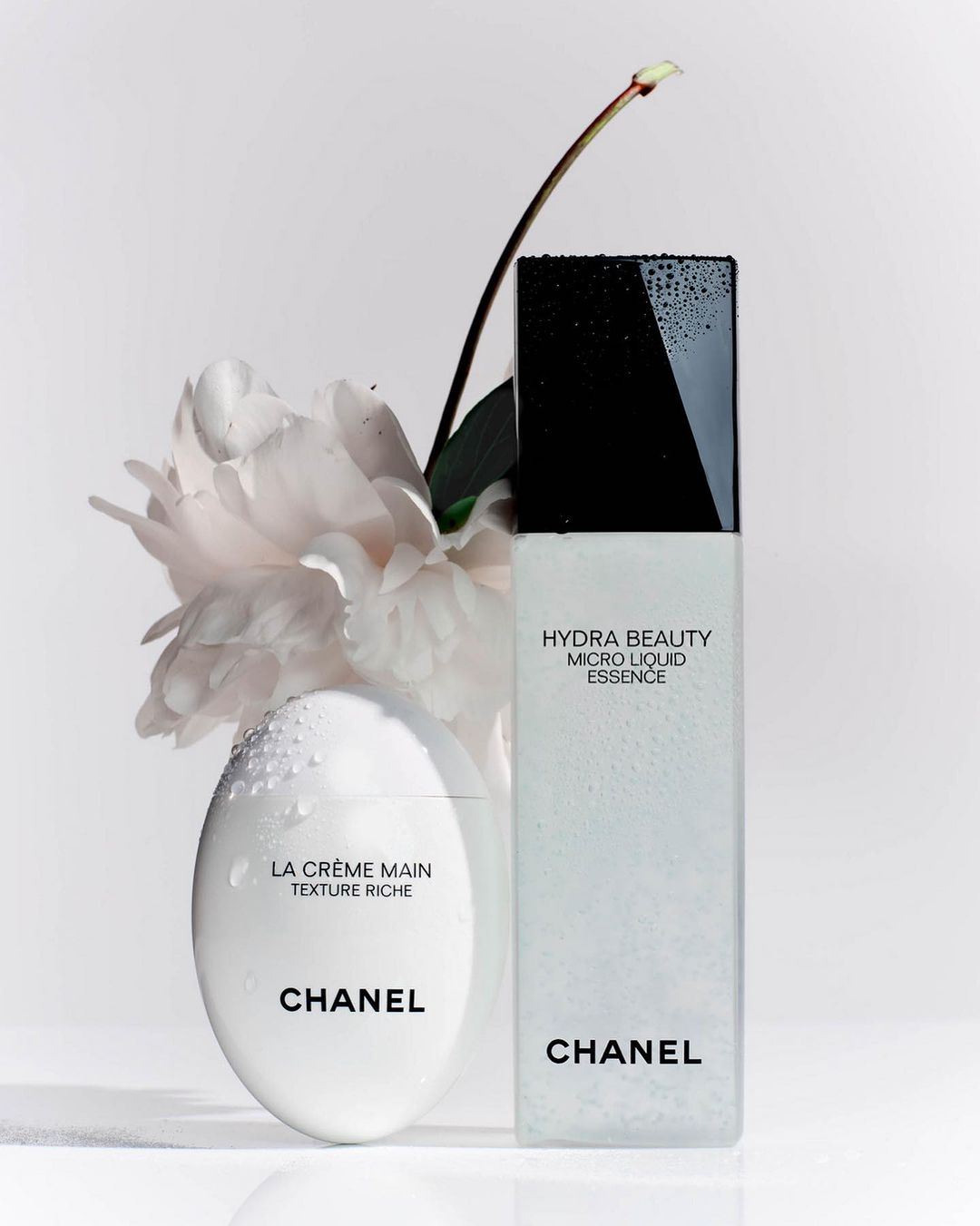 Close-up studio shot of Chanel skincare products: La Crème Main and Hydra Beauty Micro Liquid Essence, accented with a delicate white peony.