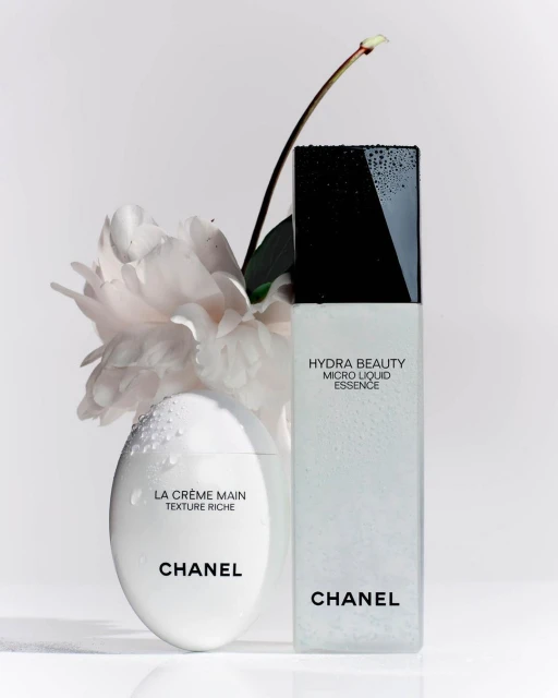 Close-up studio shot of Chanel skincare products: La Crème Main and Hydra Beauty Micro Liquid Essence, accented with a delicate white peony.