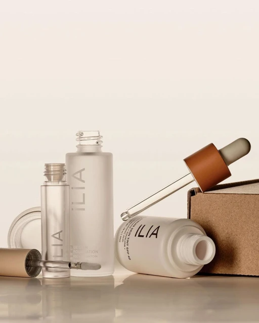 Still life of Ilia beauty products including foundation, serum and applicator with neutral color palette against white background.