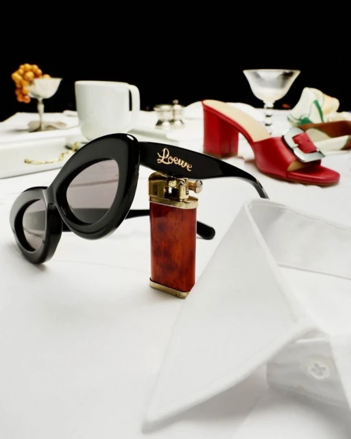 Fashion still life featuring sunglasses, a lighter, red heeled shoes, and a white dress shirt on a white surface against a black background.
