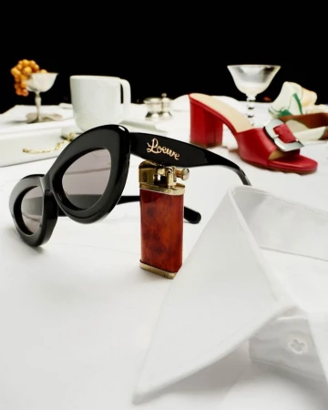 Fashion still life featuring sunglasses, a lighter, red heeled shoes, and a white dress shirt on a white surface against a black background.