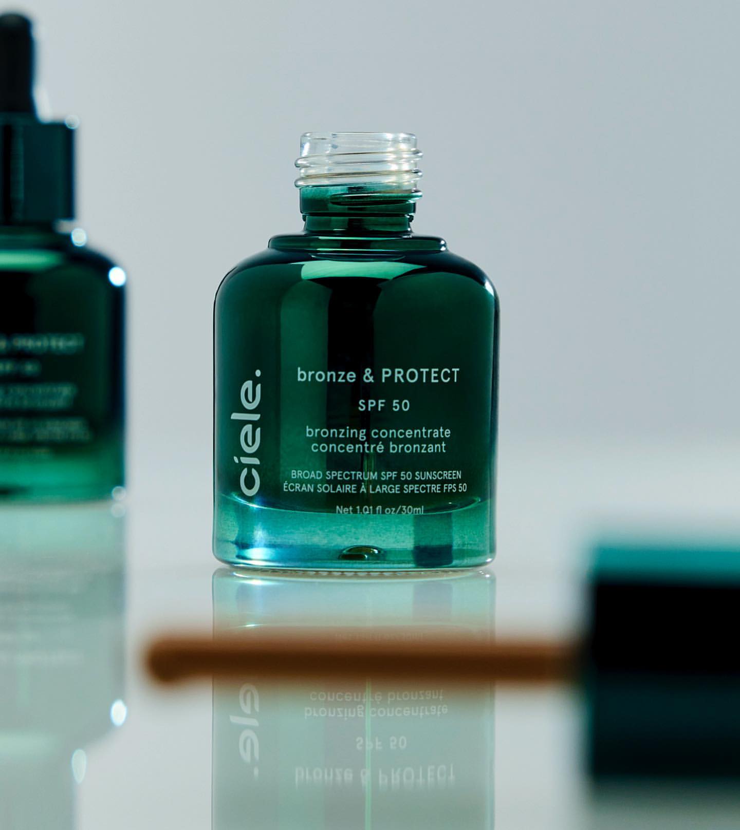 A close-up of a dark green glass bottle labeled 'bronze & PROTECT SPF 50' alongside a bottle cap and applicator, all on a white surface.