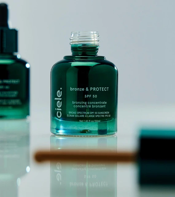 A close-up of a dark green glass bottle labeled 'bronze & PROTECT SPF 50' alongside a bottle cap and applicator, all on a white surface.