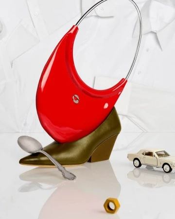 Still life studio shot of a red purse balanced on a high-heeled shoe, with a car toy, spoon, nut, and white collared shirts in the background.