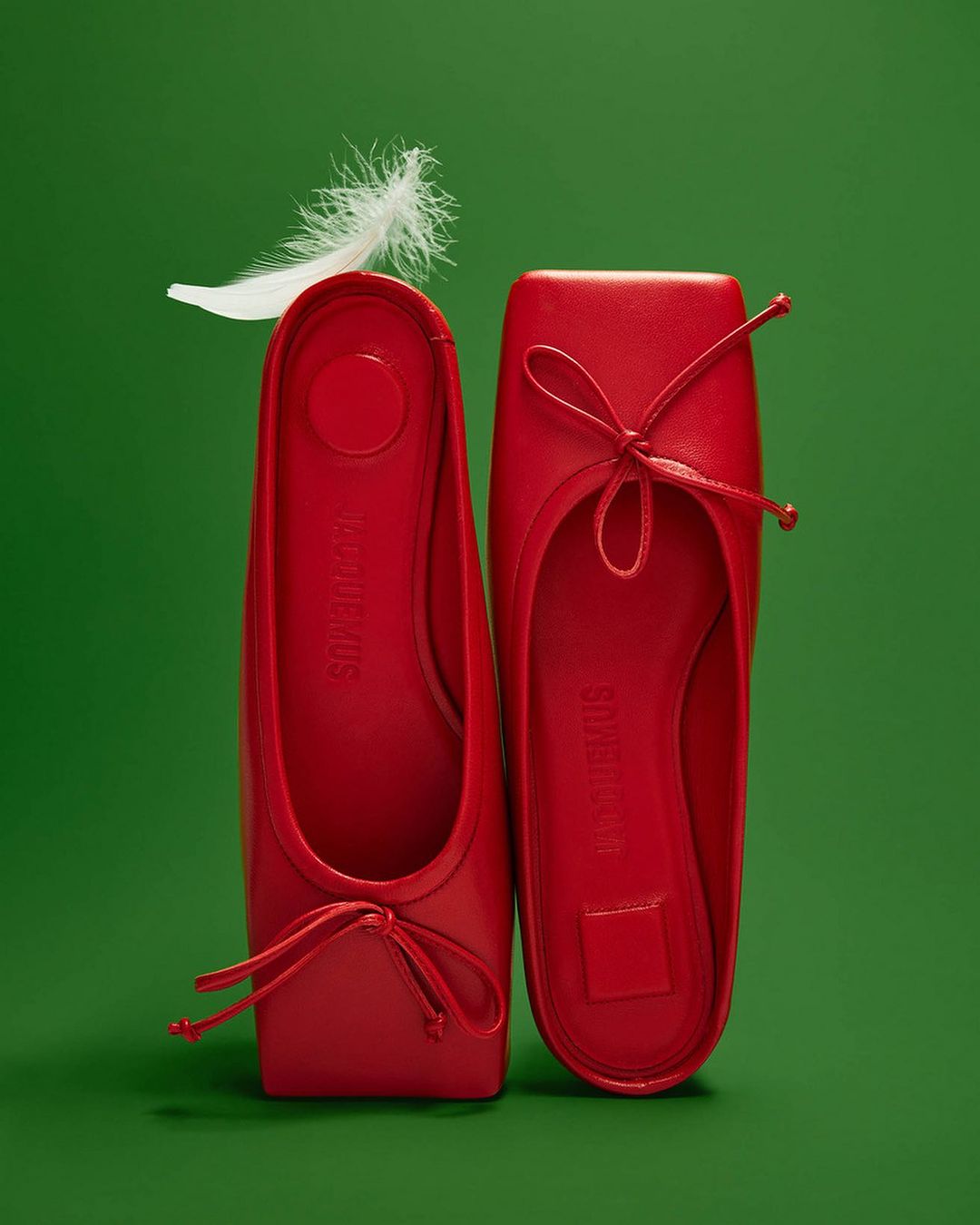 Two bright red ballet flat shoes displayed on a green background, accented with a delicate white feather for a minimalist fashion aesthetic.