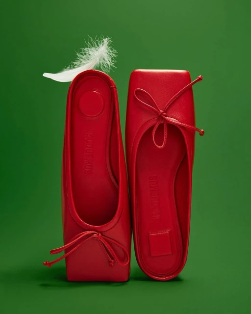 Two bright red ballet flat shoes displayed on a green background, accented with a delicate white feather for a minimalist fashion aesthetic.