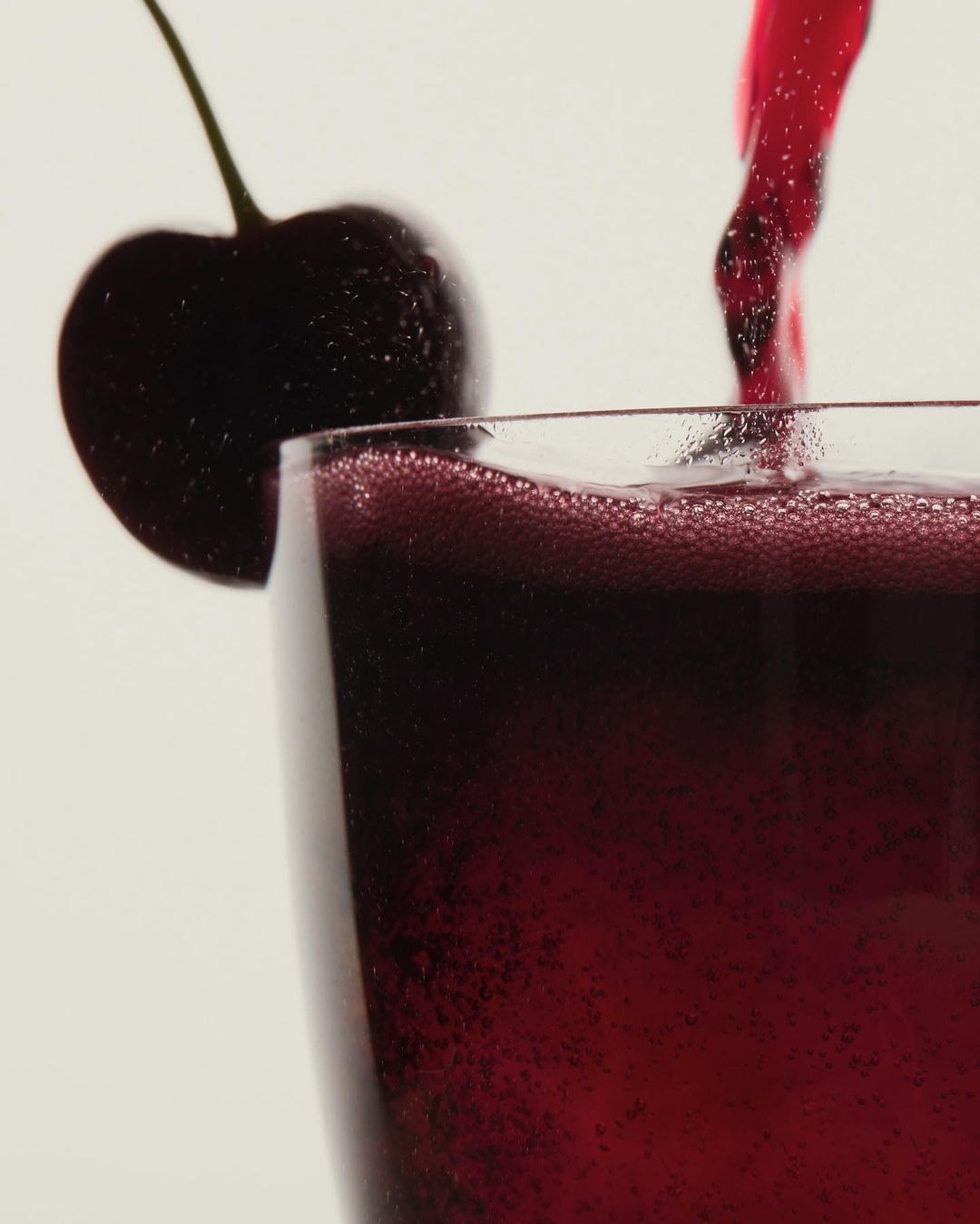 Close-up of a glass filled with a dark red beverage, garnished with a fresh cherry against a soft white background.