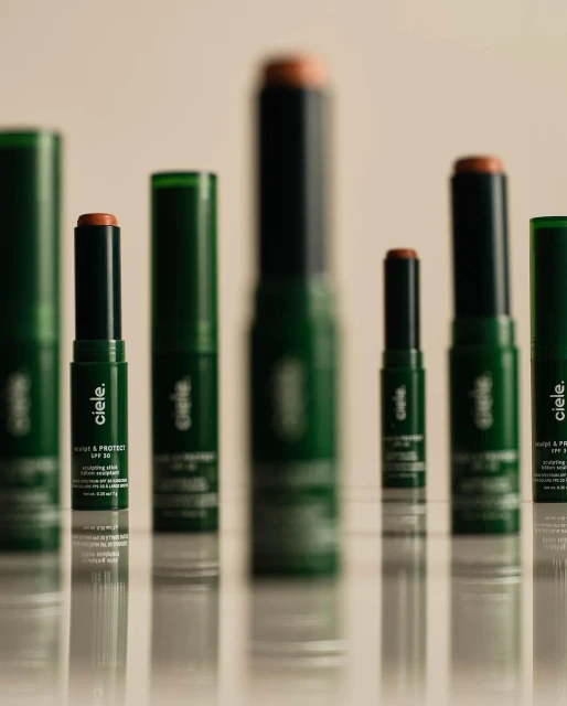 Several cie le Conceal + Protect SPF 30 makeup sticks and tubes, in dark green packaging, lined up on a reflective surface.