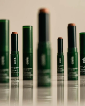 Several cie le Conceal + Protect SPF 30 makeup sticks and tubes, in dark green packaging, lined up on a reflective surface.