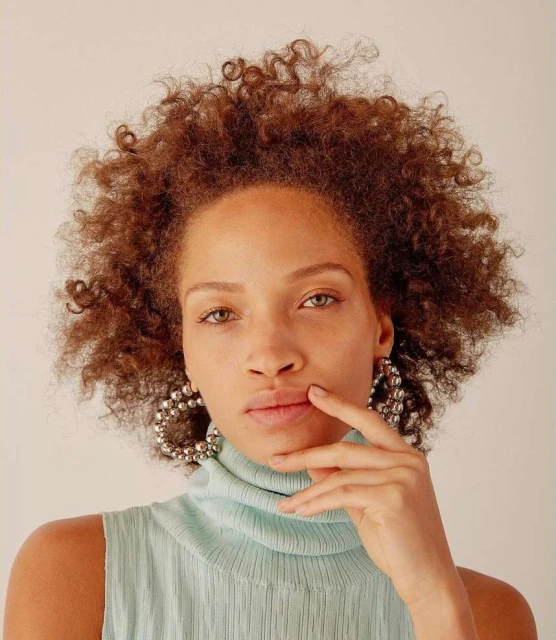 Close-up portrait of a woman with curly brown hair, wearing ball-style hoop earrings and a light blue, textured, high-neck sleeveless top.