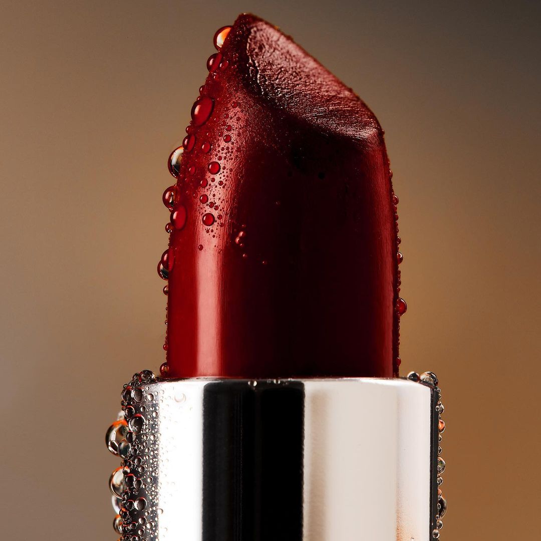 Macro shot of a deep red lipstick bullet with water droplets on a reflective silver base against a gradient brown background.