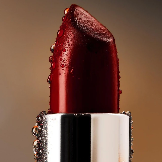 Macro shot of a deep red lipstick bullet with water droplets on a reflective silver base against a gradient brown background.