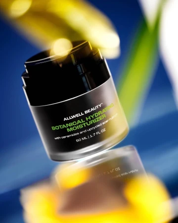 A black cosmetic jar of Allwell Beauty botanical hydrating moisturizer centered against a blue background with blurred yellow foreground elements