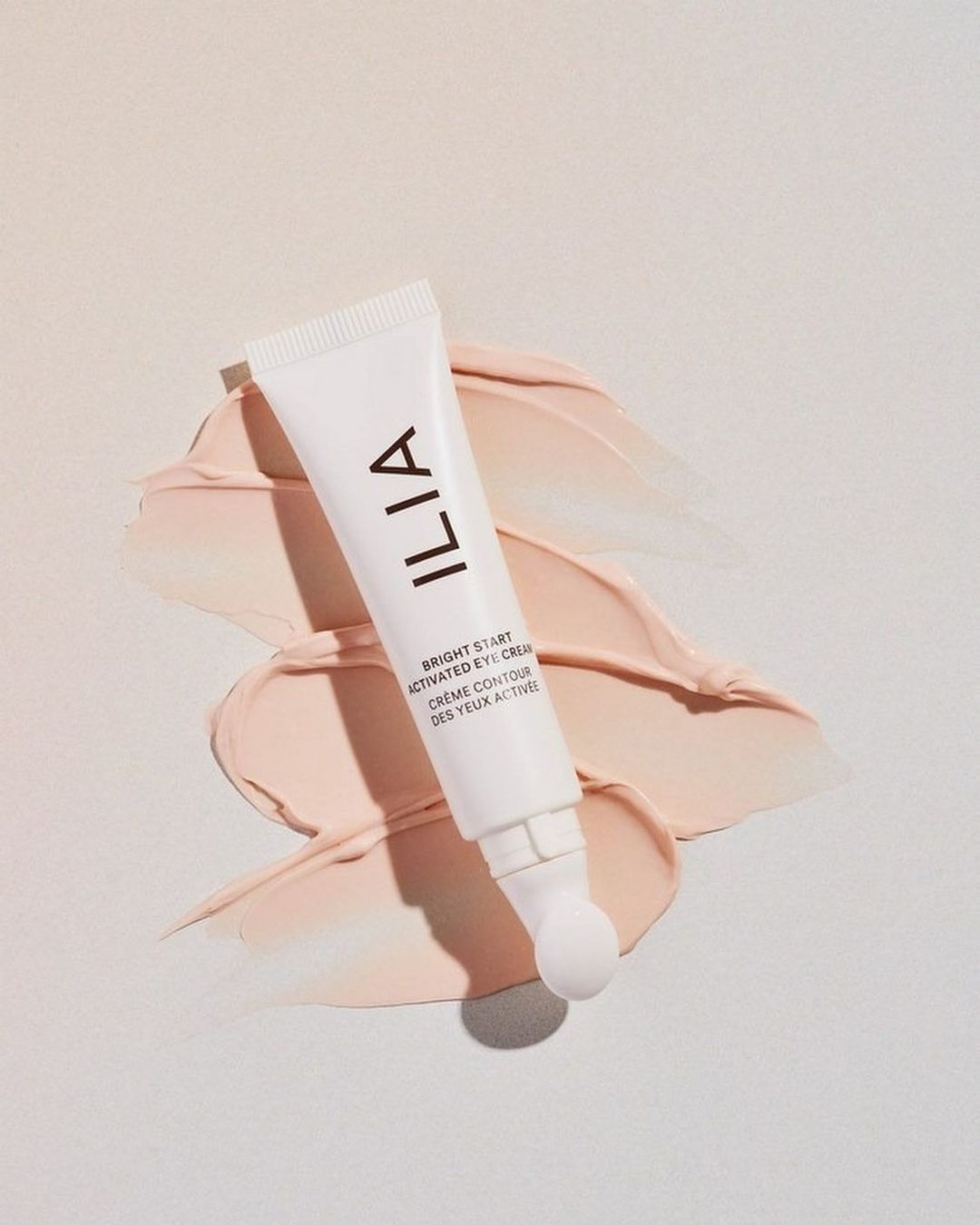 Close-up of a white tube of Ilia eye cream lying on a smear of peach-colored cream on a white background, showcasing the product and texture.