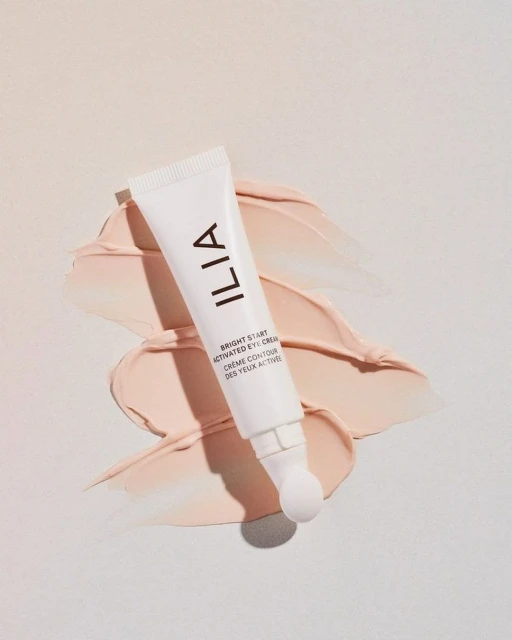 Close-up of a white tube of Ilia eye cream lying on a smear of peach-colored cream on a white background, showcasing the product and texture.