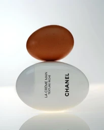 Brown egg resting balanced on top of a white oval Chanel hand cream tube against a bright light background