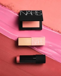NARS cosmetics products including a blush compact and cream stick arranged on a pink and coral textured background.