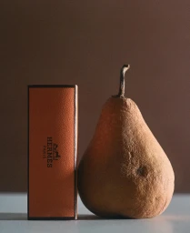 An orange textured Hermes box standing vertically next to a ripe brown pear on a light surface.