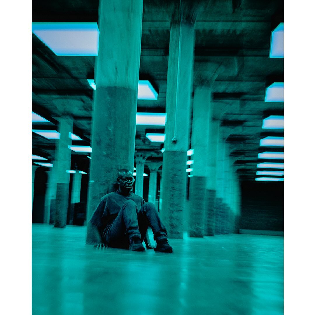 Person in dark shirt and jeans sits on the floor in a hall with concrete pillars and blue light fixtures, low angle perspective.