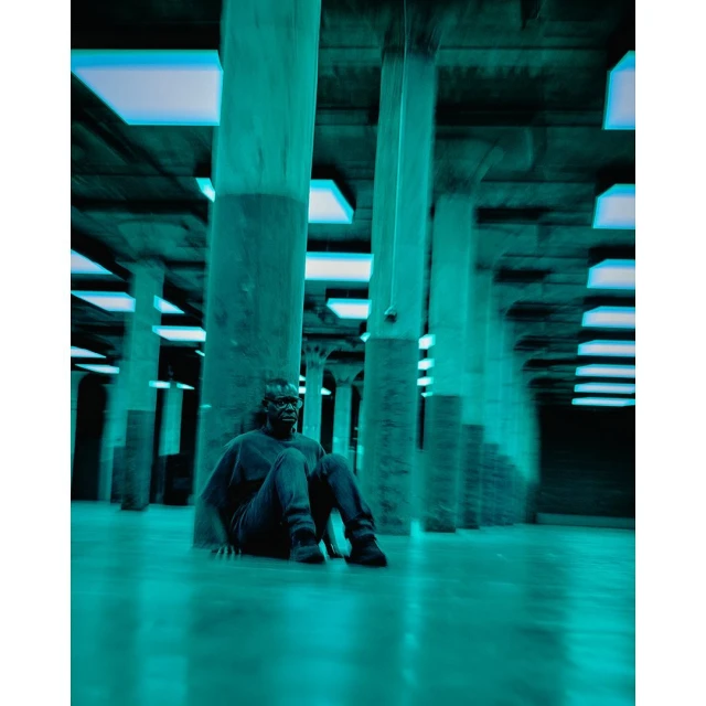 Person in dark shirt and jeans sits on the floor in a hall with concrete pillars and blue light fixtures, low angle perspective.