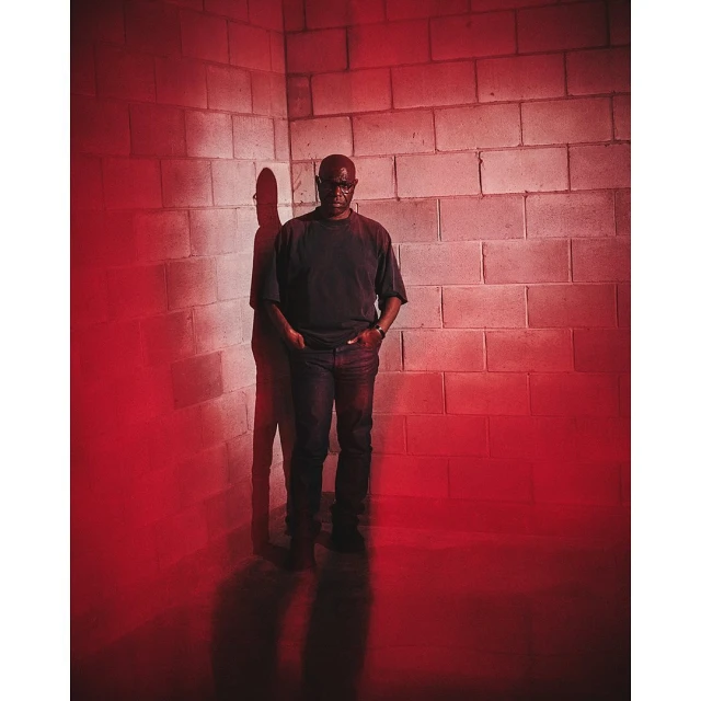 Portrait of a Black man standing in front of a cinder block wall, cast in dramatic red lighting with a noticeable shadow. He wears casual clothes.